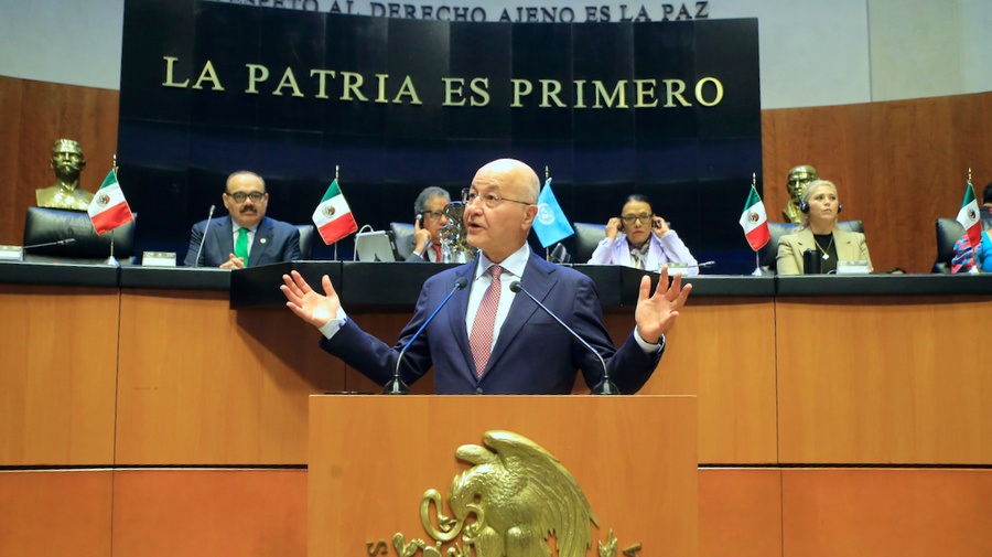 A man gestures while speaking behind a lectern with a row of people looking on from a raised platform behind