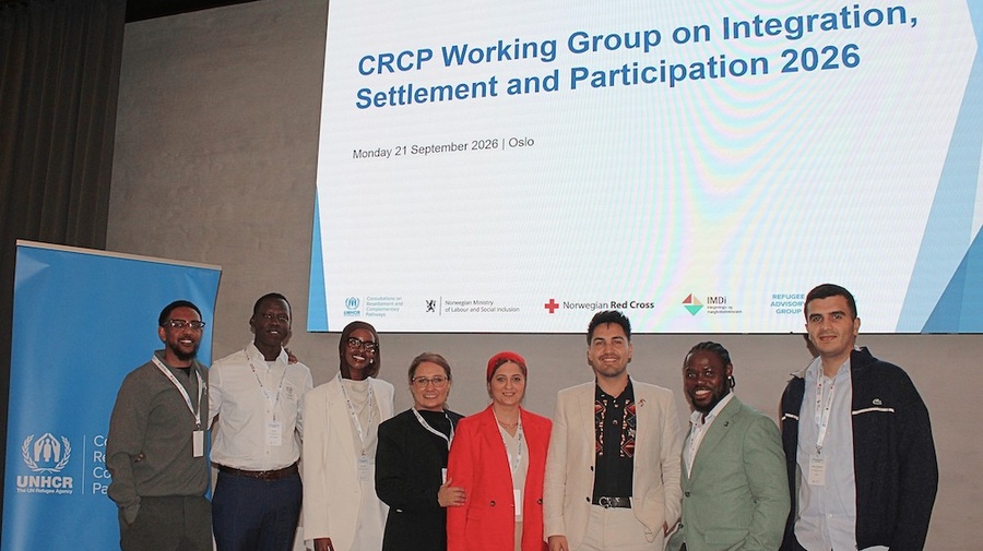 Eight members of the Refugee Advisory Group stand on stage in front of a screen reading "CRCP Working Group on Integration, Settlement and Participation 2026."