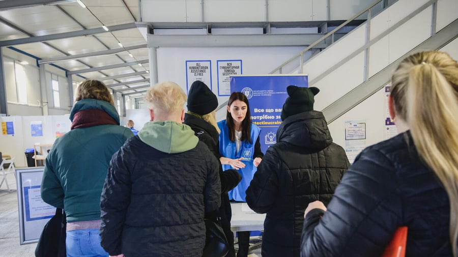 Pictured from behind, 5 people queue at a desk where a UNHCR staff member assists. 