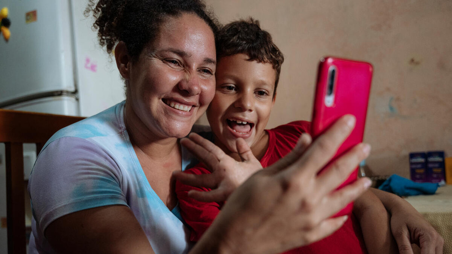 A woman and a boy smile excitedly, looking at a mobile phone they're holding up in front of them. The boy is waving.