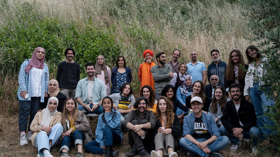 A large group of people are pictured, smiling, outdoors in front of tall grasses and bushes