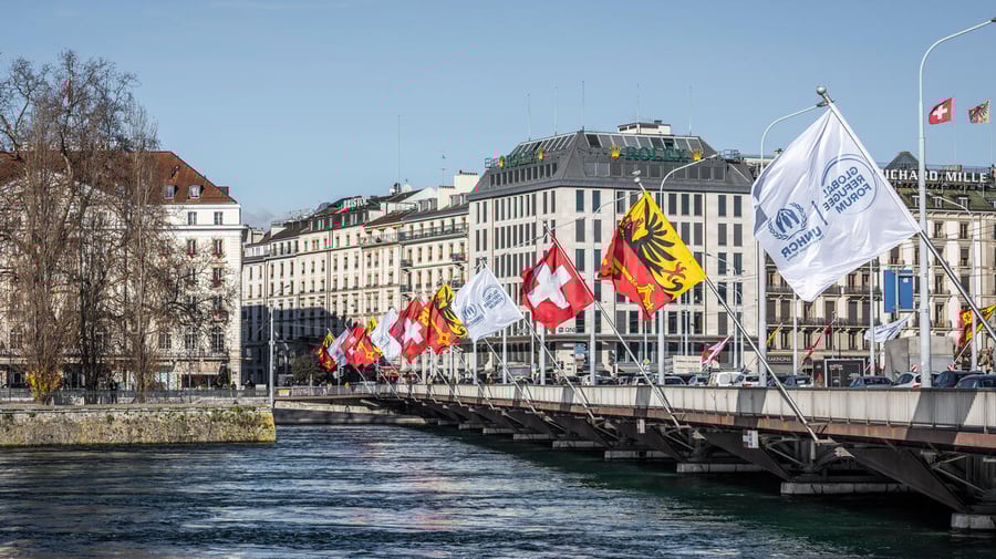 Flags fly along the length of a bridge over a wide river with buildings in the background