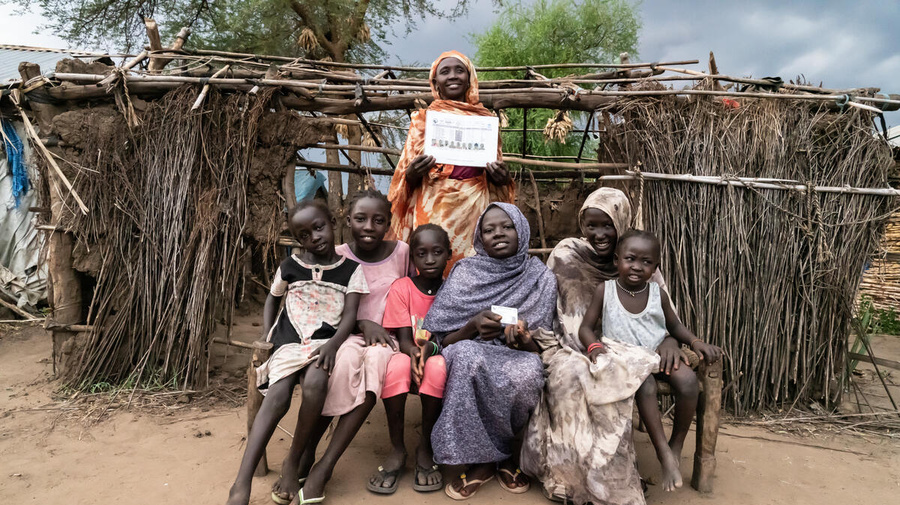 A family is pictured, smiling, in front of a shelter, with two members displaying their IDs.