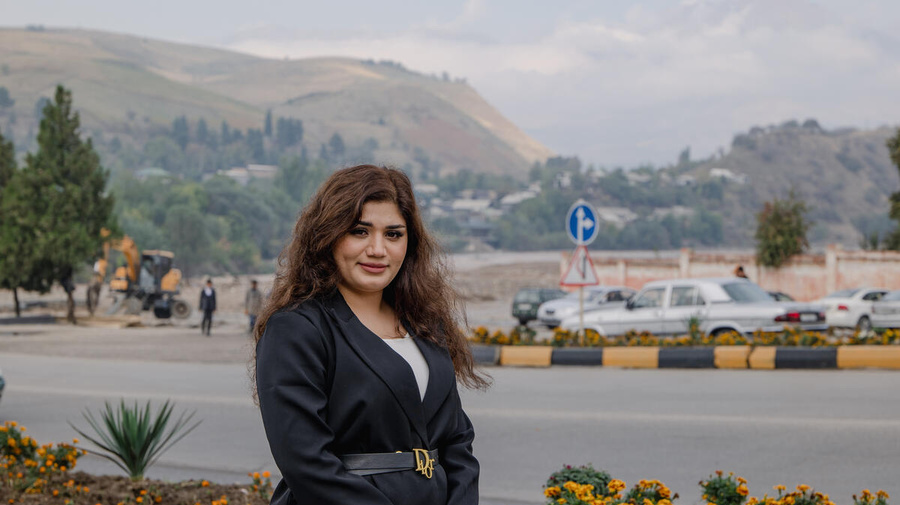 A smiling woman pictured in front of flowers alongside a road, with large hills in the distance.