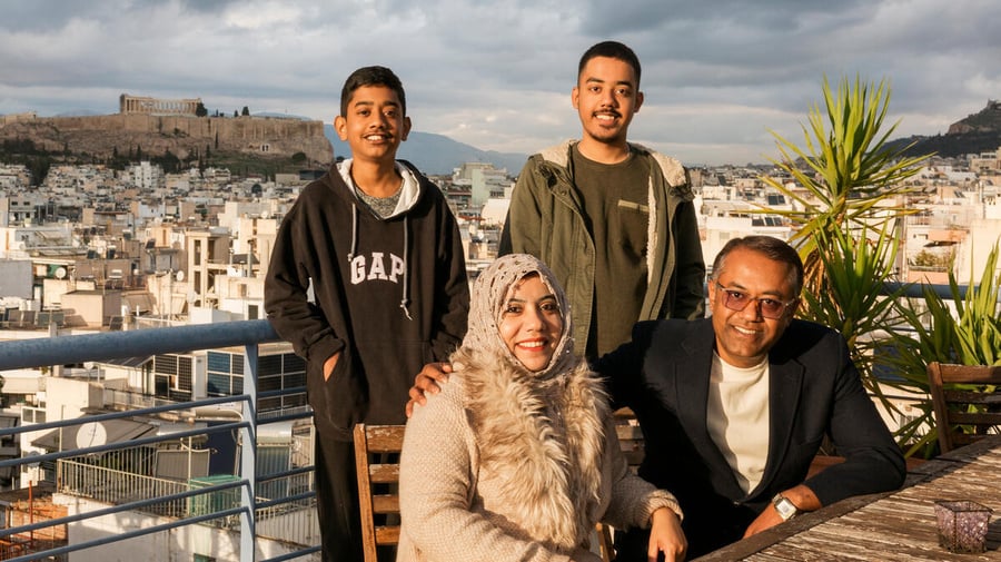 A middle-aged couple sit on a balcony with their two sons standing behind them, with a view of Athens behind them. 