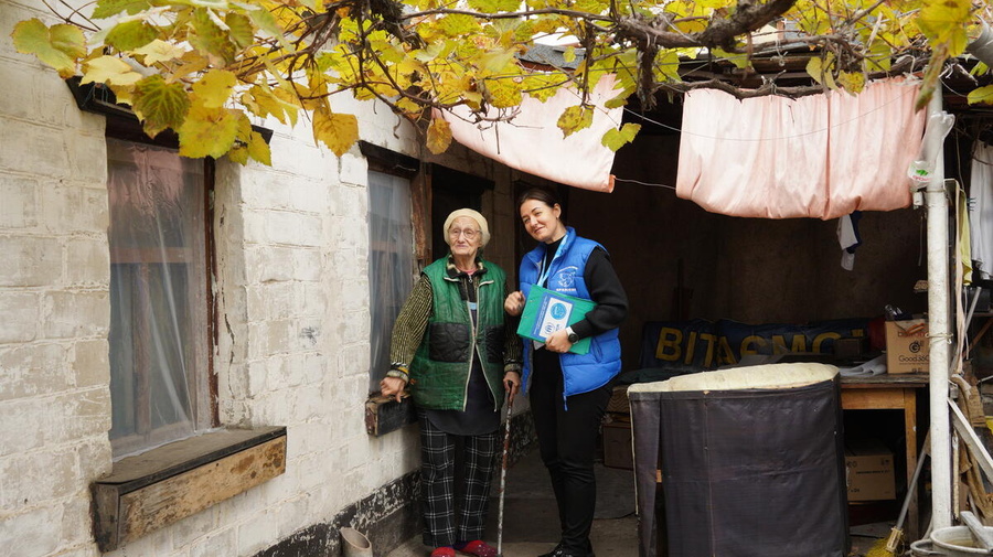 A UNHCR staff member and a woman in her 80s stand outside a home, under the branches of a tree