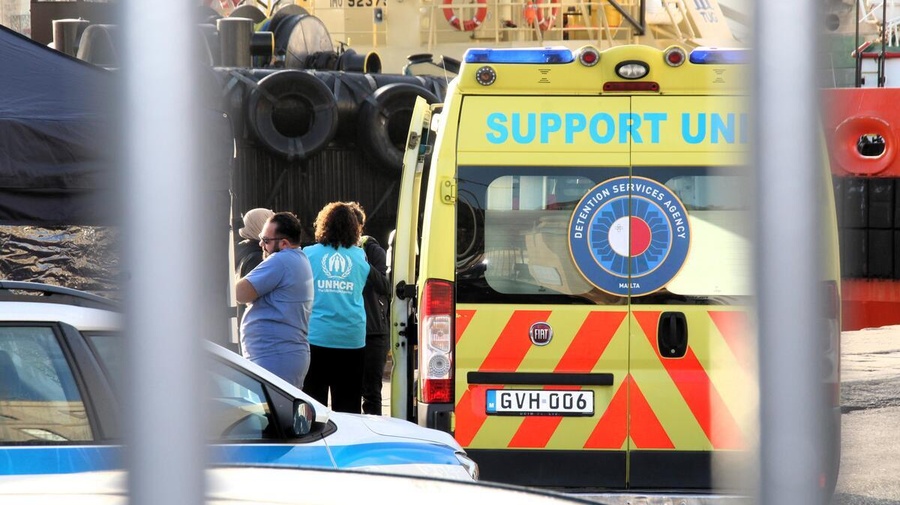 UNHCR staff stand beside a detention services agency vehicle parked next to a port.