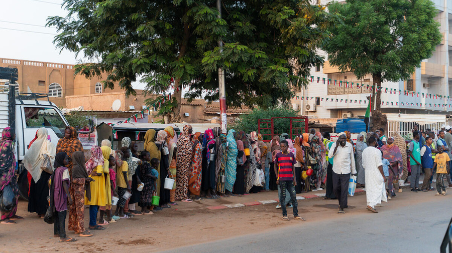 Families from El Fasher queue outside along a street.
