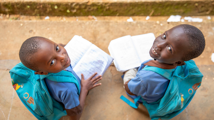 Two girls, 7- and 8-years-old, from Central African Republic smile up at the camera from where they're sitting outside, wearing matching 'Education cannot wait' backpacks and holding open books.