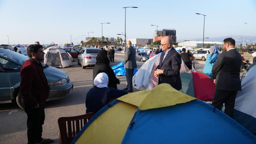 A man in a suit speaks to a family in an open area filled with tents and vehicles 