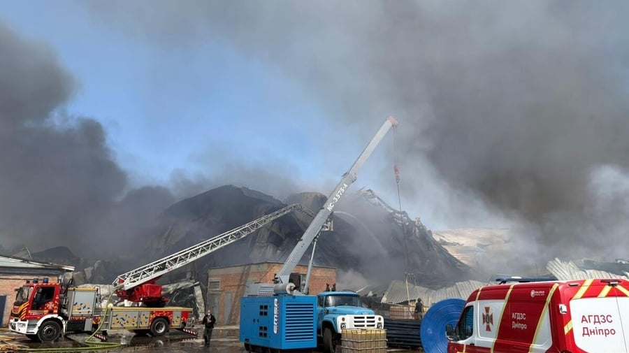 Black smoke surrounds a damaged warehouse, with emergency vehicles attending