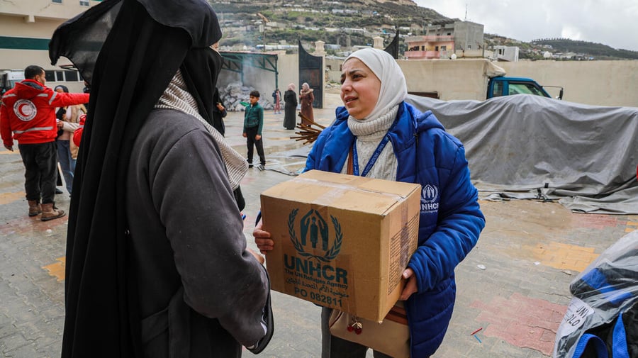 A woman in a UNHCR jacket hands a box of UNHCR supplies to another woman