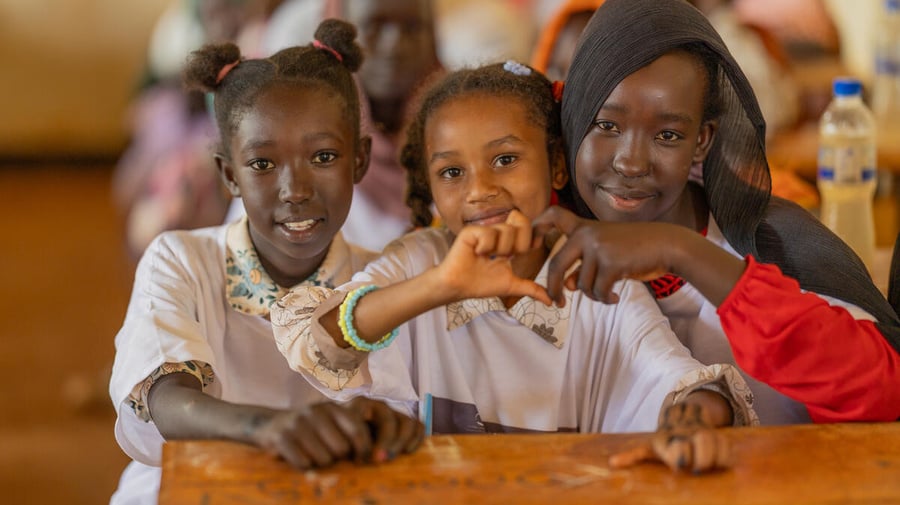Three Sudanese refugee pupils sit at a desk, smiling, two of them holding their hands together to make a heart shape.