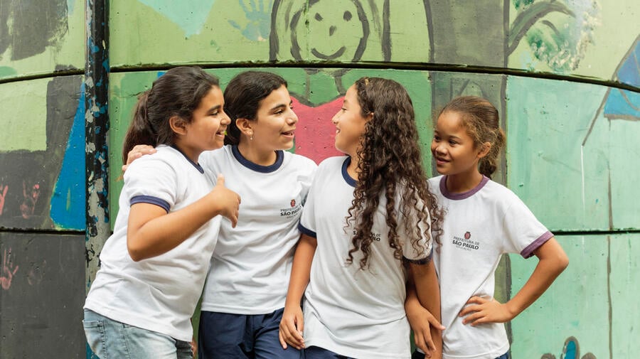 UNHCR UN Refugee Agency - How we do resettlement - ALT text: Four young girls enjoy chatting to each other while standing outside their classroom at the Duque de Caxias Municipal School in the Glicerio neighborhood of downtown Sao Paulo in Brazil. 