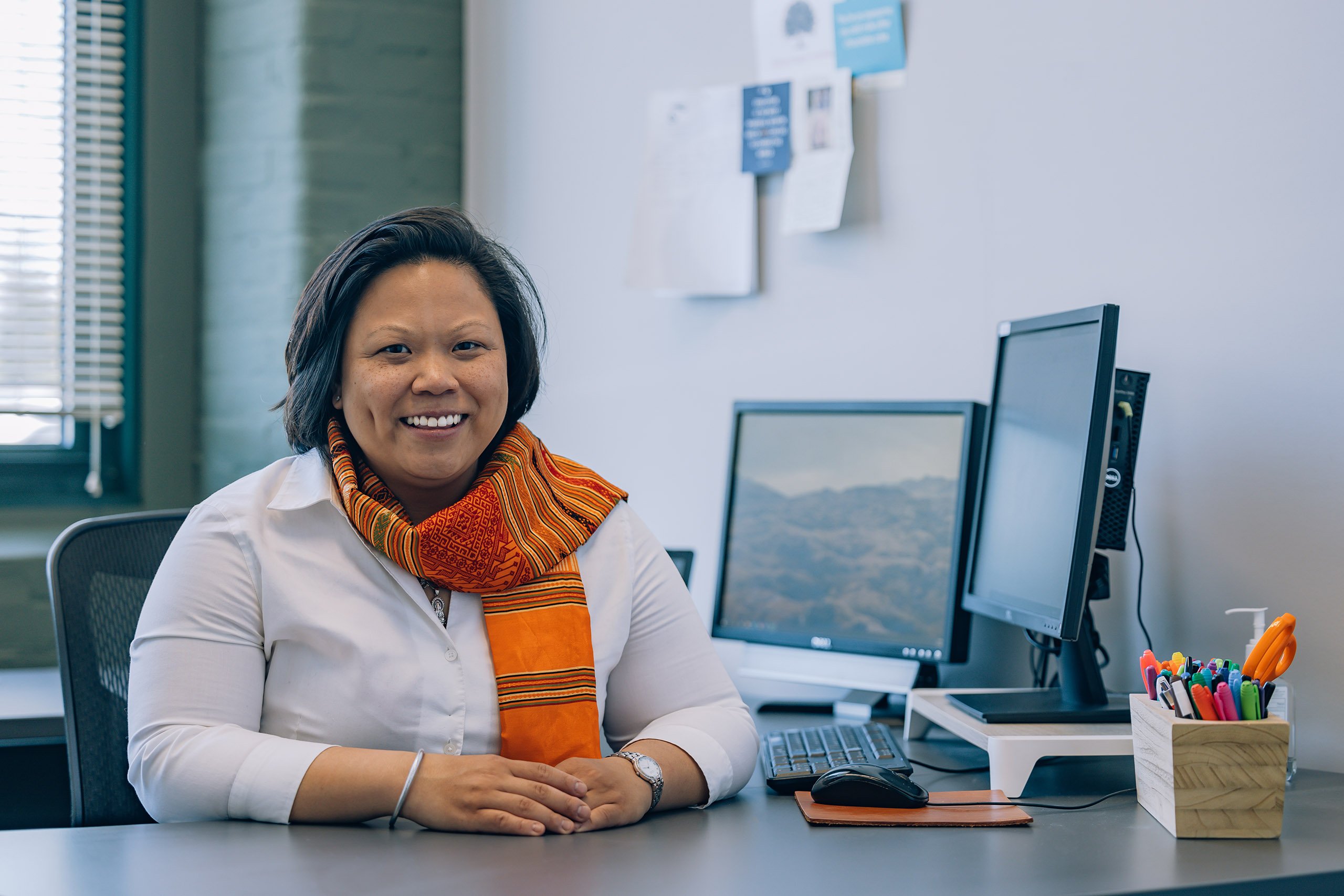 A Laotian-American woman wearing a traditional orange scarf sits behind an office desk next to a computer.