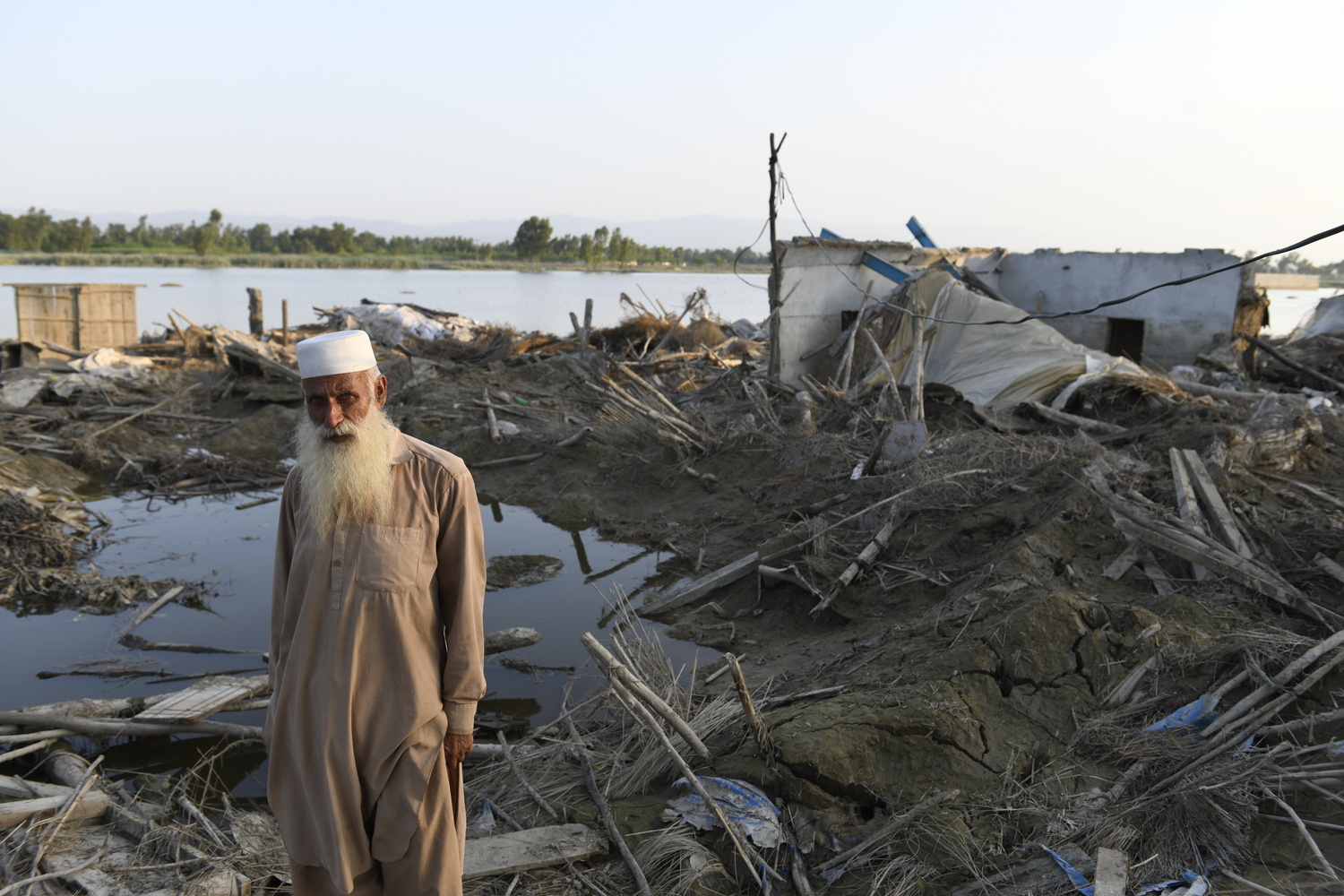 An elderly man stands in front of a house destroyed by flooding.