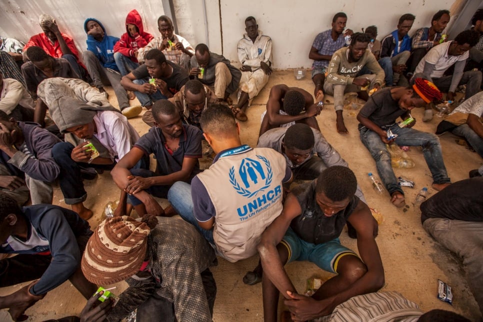 A group of disembarked refugees and migrants sit on the floor, joined by a UNHCR staff member wearing a tan UNHCR vest with blue logo.
