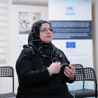 A person speaks in a community center, using hand gestures while sharing their experience.