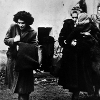 Black-and-white historical photograph showing four people outdoors near a plain building wall. One person in the foreground walks while carrying a large bag over a shoulder. Three other people stand close together behind, wearing long coats and head coverings. The ground appears muddy and scattered with debris, and the image has a grainy, documentary-style appearance.