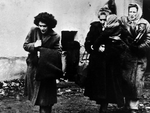 Black-and-white historical photograph showing four people outdoors near a plain building wall. One person in the foreground walks while carrying a large bag over a shoulder. Three other people stand close together behind, wearing long coats and head coverings. The ground appears muddy and scattered with debris, and the image has a grainy, documentary-style appearance.