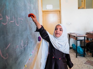 An Afghan girl holds up her arm towards a blackboard in a classroom, as others sit at desks behind her.