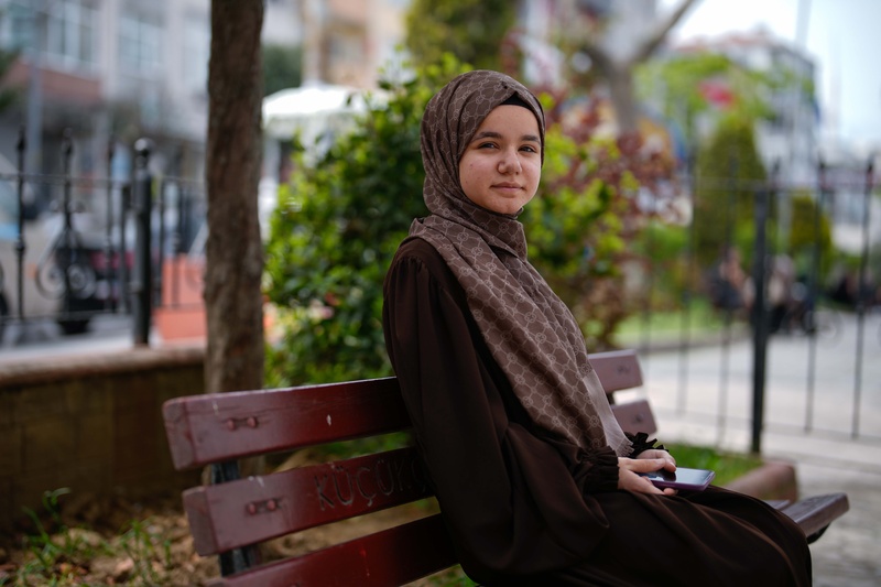 A person sits on a park bench holding a smartphone, with trees and an urban streetscape in the background.