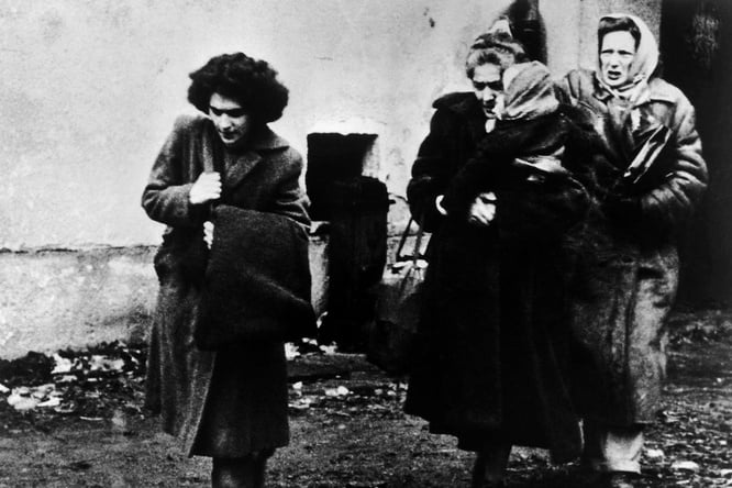 Black-and-white historical photograph showing four people outdoors near a plain building wall. One person in the foreground walks while carrying a large bag over a shoulder. Three other people stand close together behind, wearing long coats and head coverings. The ground appears muddy and scattered with debris, and the image has a grainy, documentary-style appearance.
