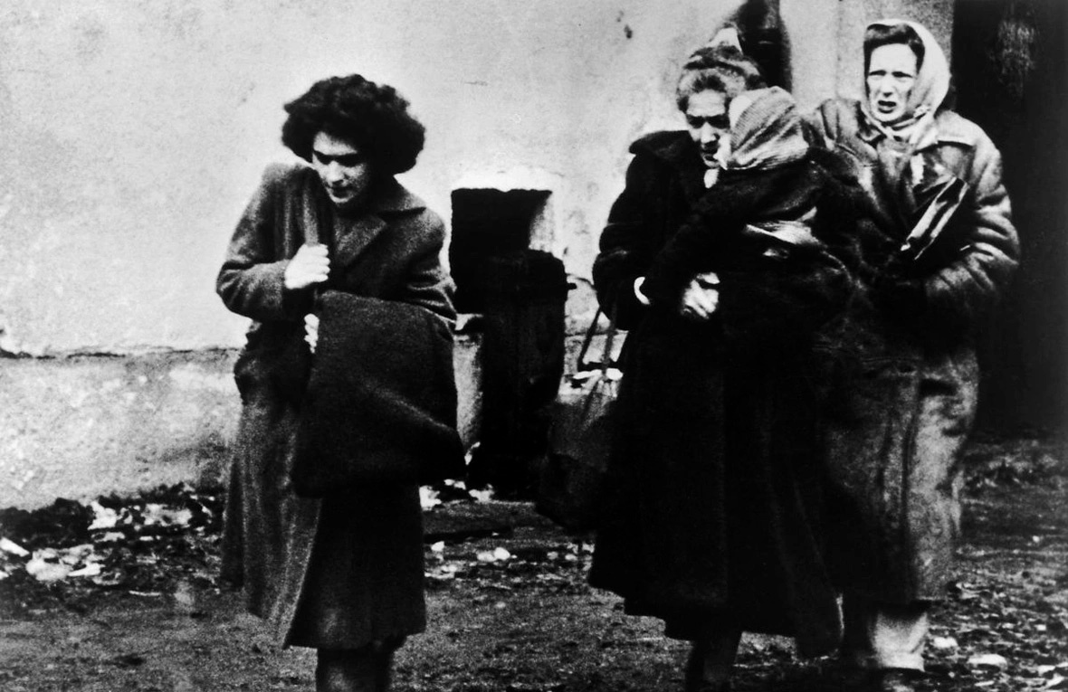 Black-and-white historical photograph showing four people outdoors near a plain building wall. One person in the foreground walks while carrying a large bag over a shoulder. Three other people stand close together behind, wearing long coats and head coverings. The ground appears muddy and scattered with debris, and the image has a grainy, documentary-style appearance.