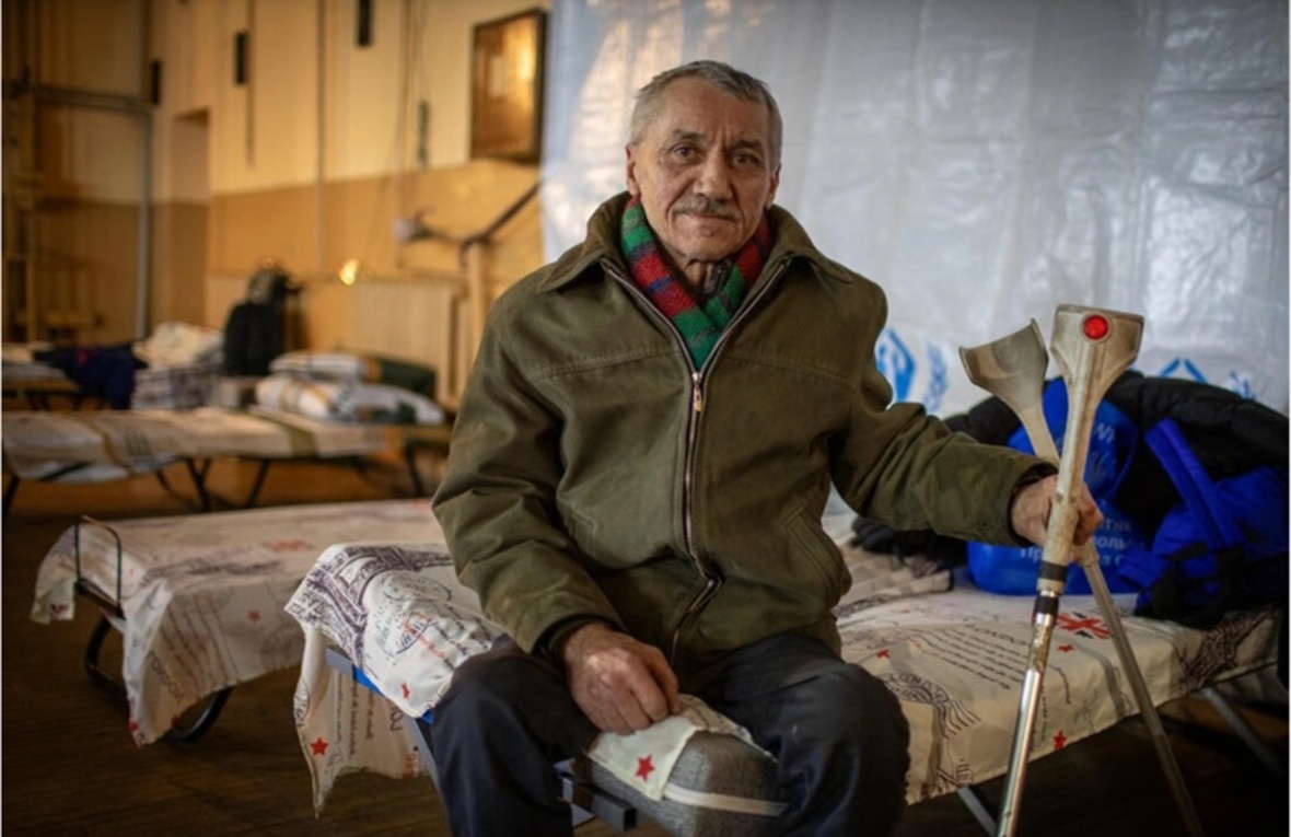 Чоловік у віці сидить на розкладному ліжку в гуртожитку, тримаючи милиці.