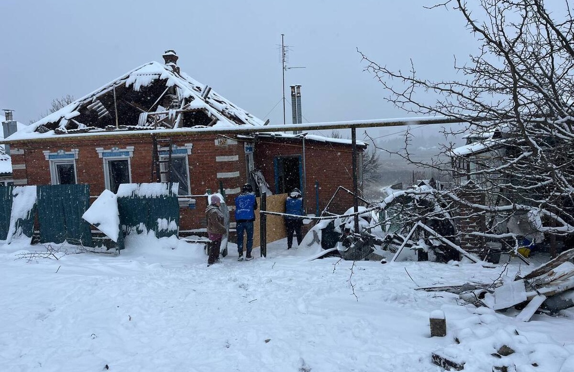 Three people stand outside in the snow, facing a damaged building with a collapsed roof