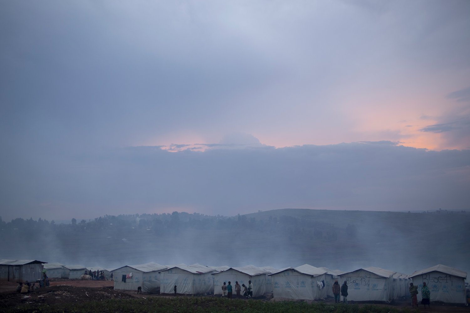 People stand near a row of white tents on a hillside at dusk.