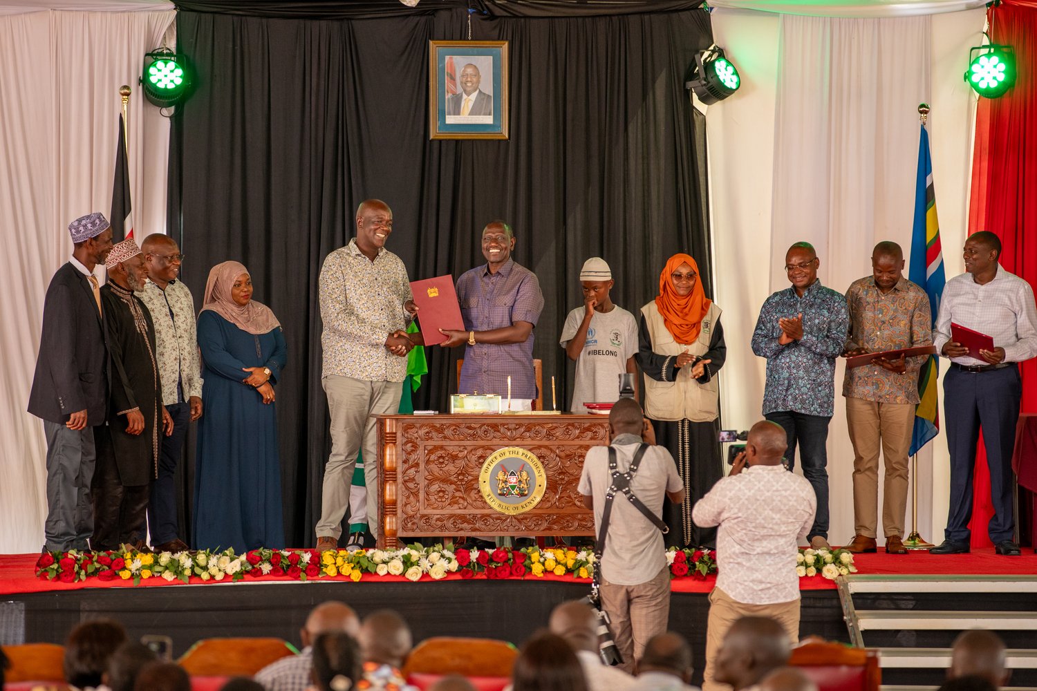 People stand on a stage during an official ceremony. A man in the centre holds up a red-bound document.