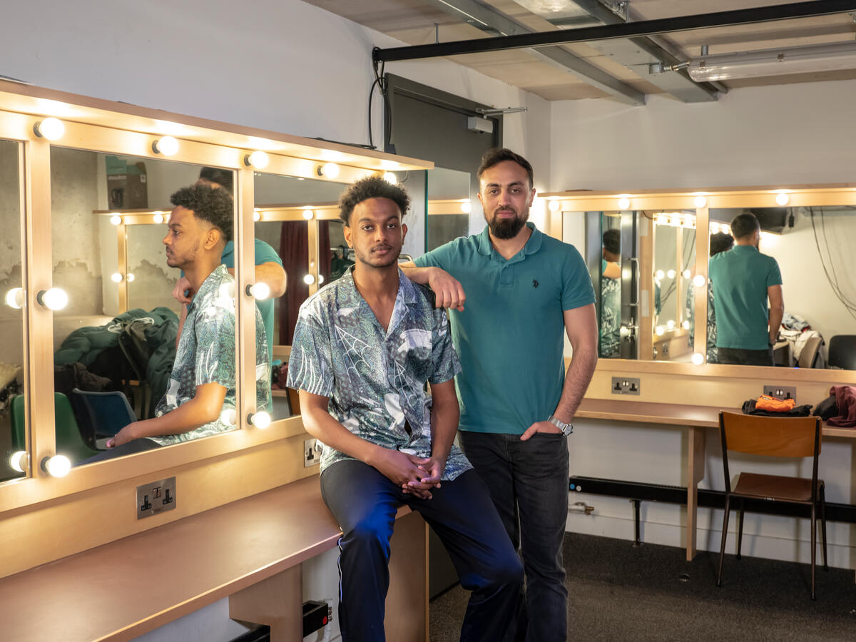 Two men stand close together looing at the camera in a theatre dressing room