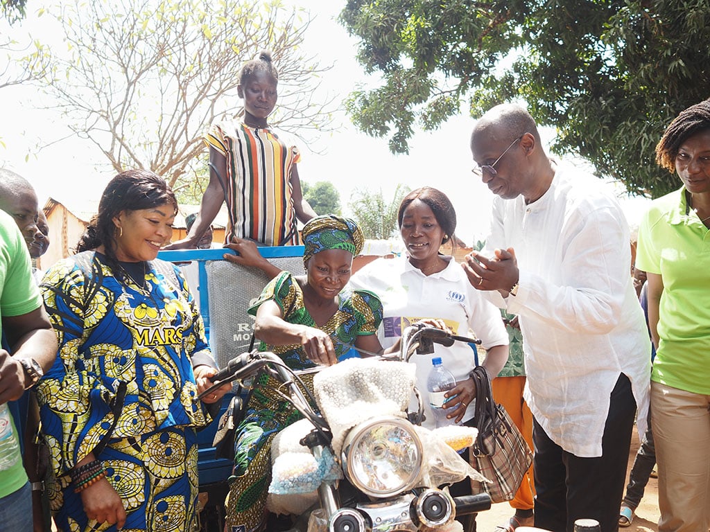 A group of people, smiling, gathers around a tricycle motorcycle