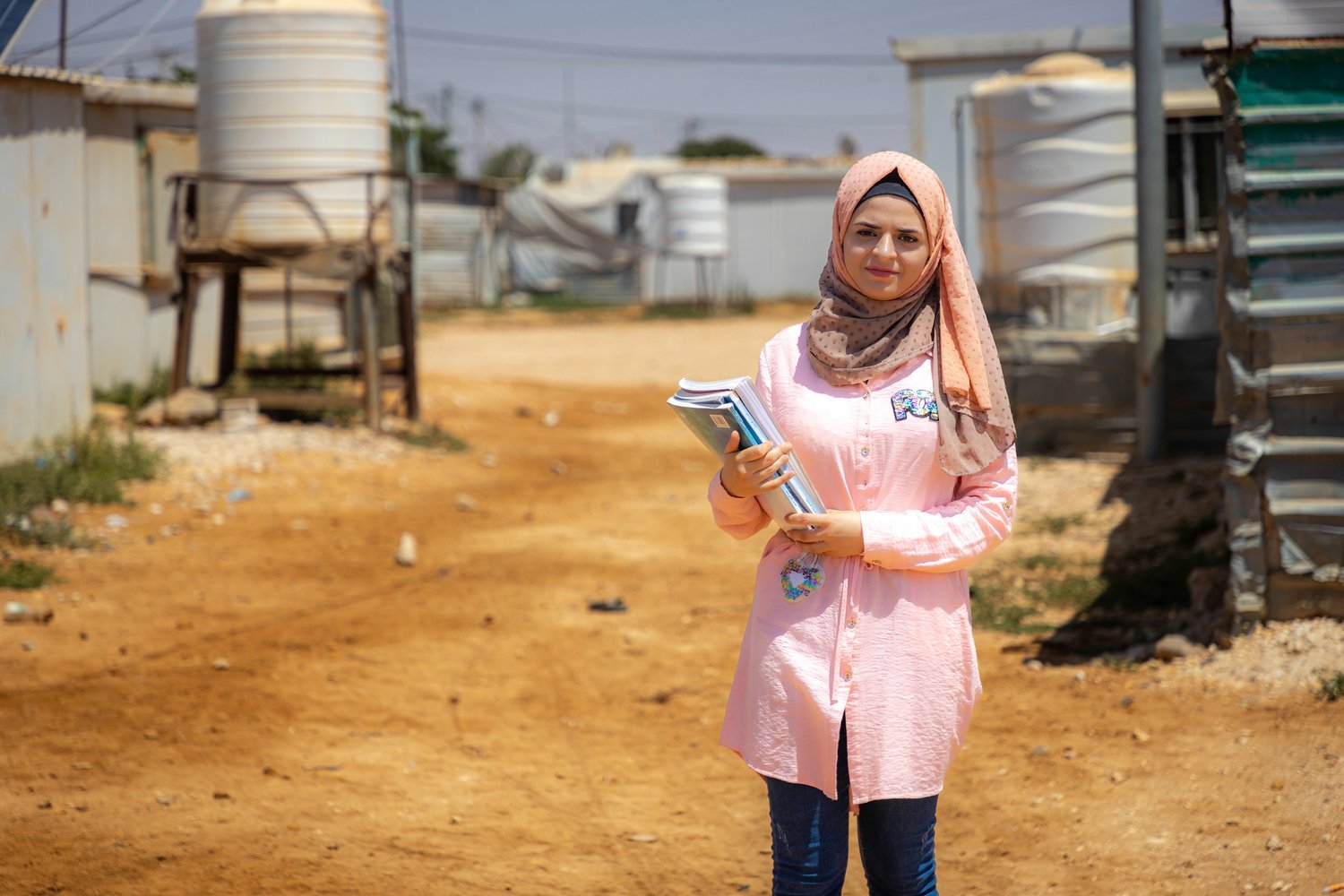 A young woman wearing a pink headscarf stands in a camp holding some school books.