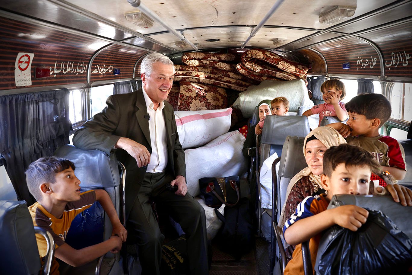 A smiling man in a suit stands inside a bus with women and children seated around him and mattresses piled in the rear of the vehicle