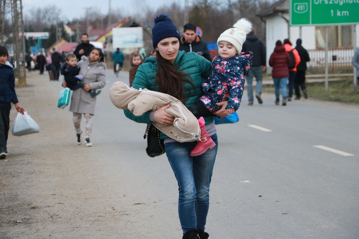 A young woman walks along the side of a road carrying a toddler and a baby in her arms