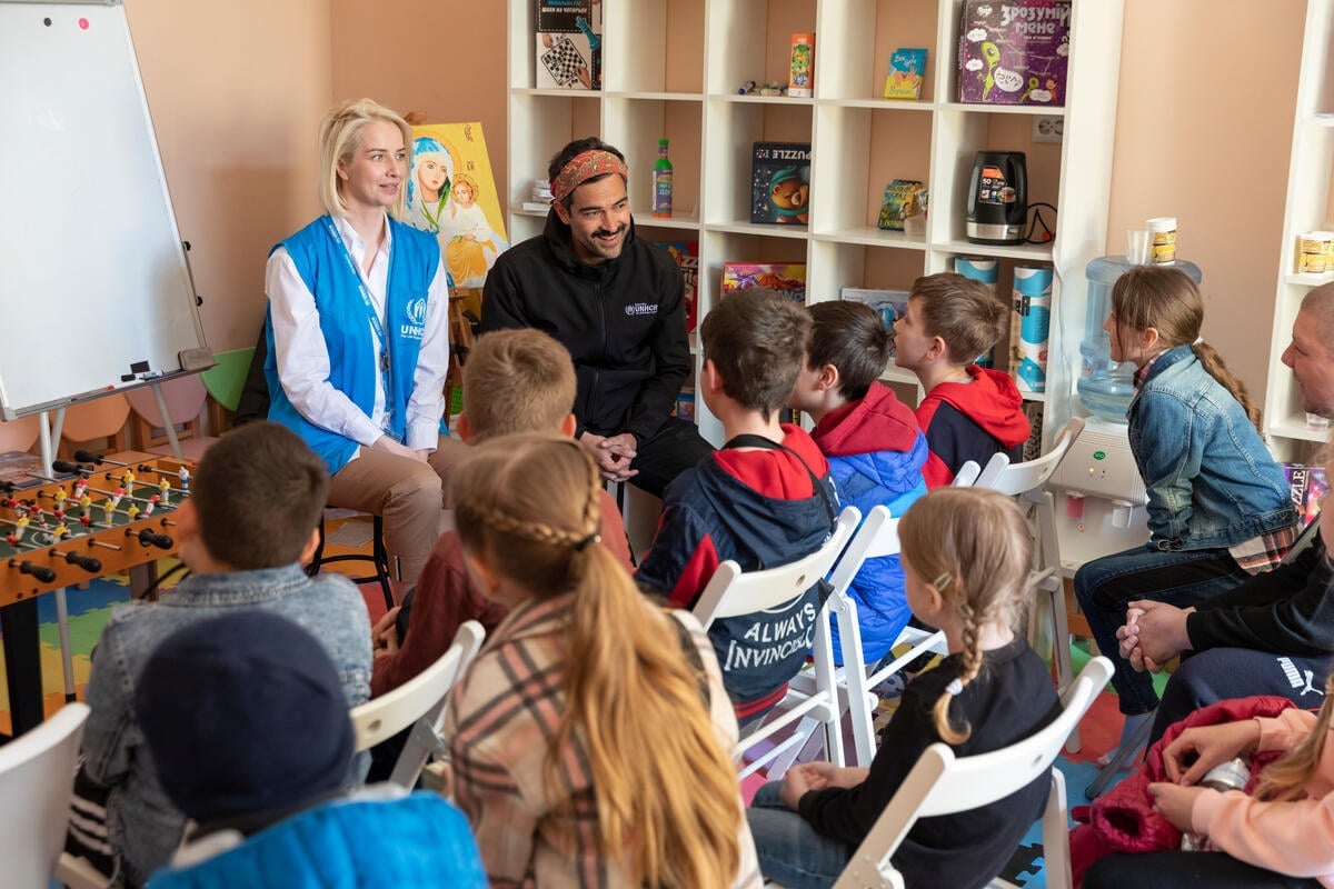 Mexican actor Alfonso Herrera and a UNHCR staff member sit in a classroom in front of rows of children.