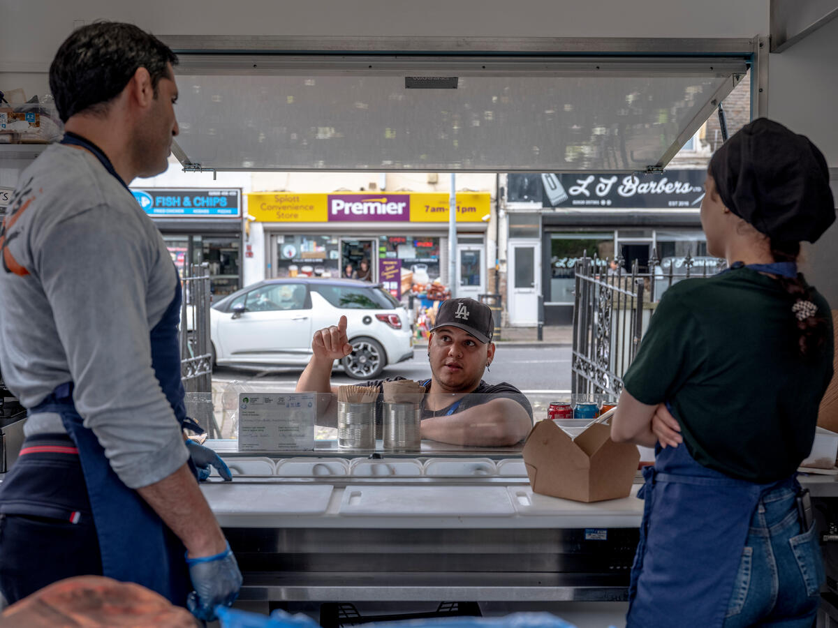 Two people inside a food truck speak with one man stood at the counter, outside the truck.