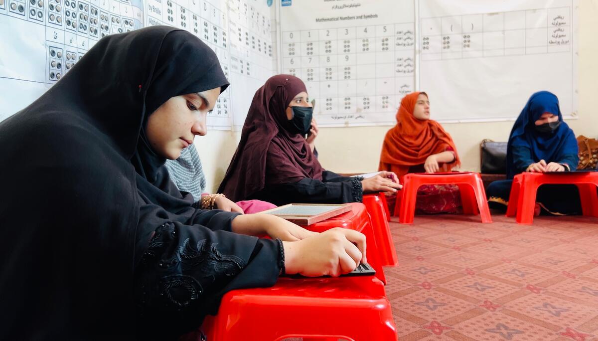 Helai Noori sits on the floor of a classroom with other women and reads Braille on a wooden tablet.