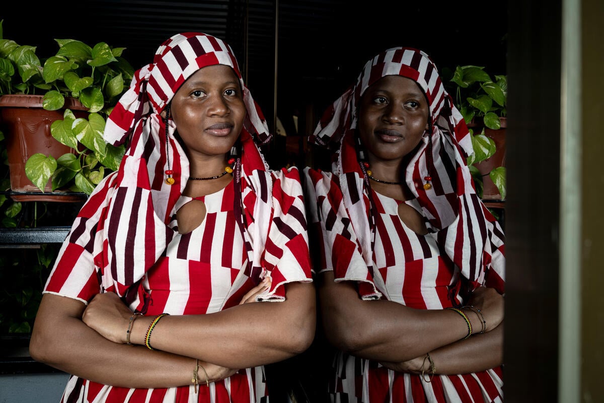 A woman in a red and white striped dress and headscarf leans against a window in which she is reflected