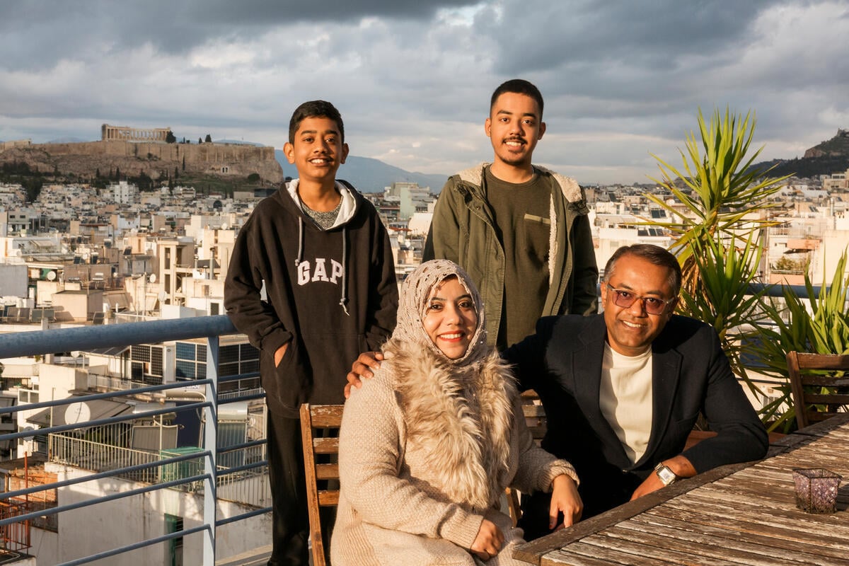 A middle-aged couple sit on a balcony with their two sons standing behind them, with a view of Athens behind them.