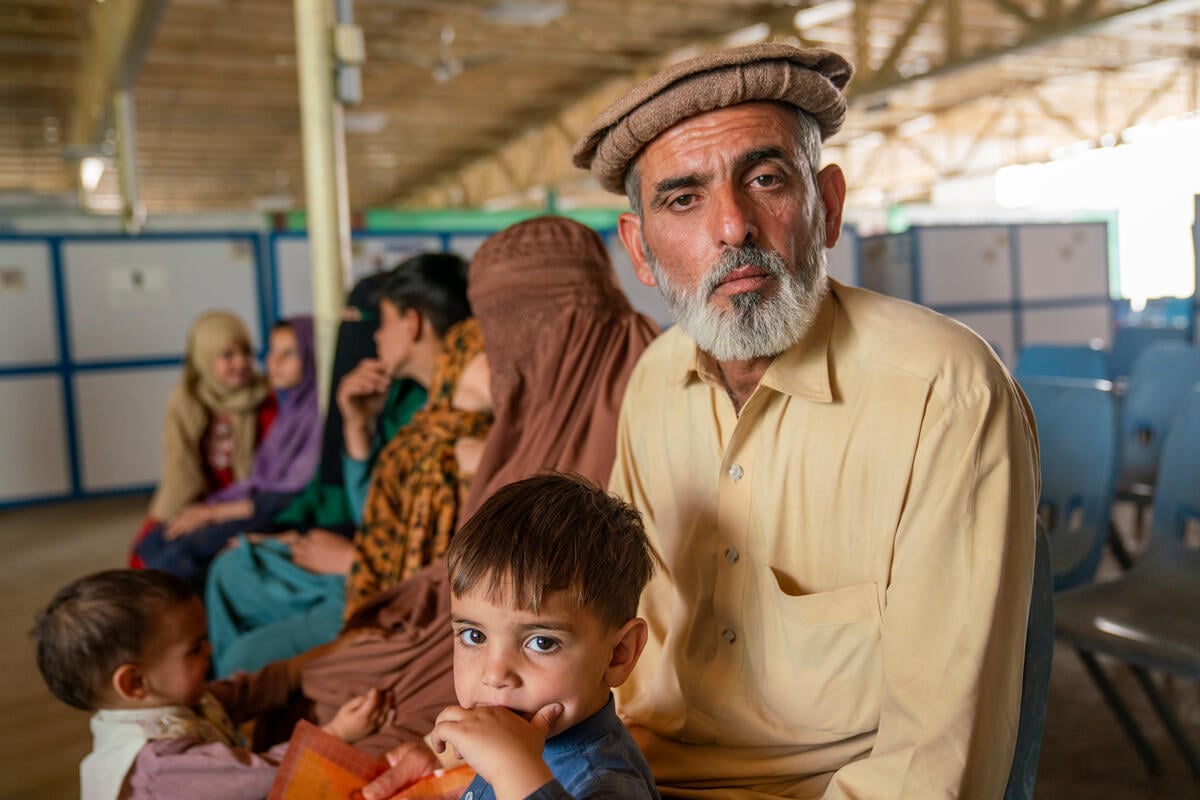 A man with a grey beard wearing a traditional pakol or rolled-up cap sits in a large, open-sided reception centre holding a young boy on his lap as other family members sit behind him