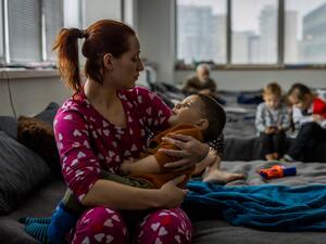A woman sits on a camp bed holding her young son in a collective shelter for refugees.
