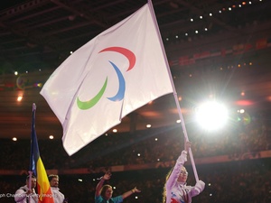 A woman wearing glasses holds a large IPC flag above her head in a brightly lit stadium.