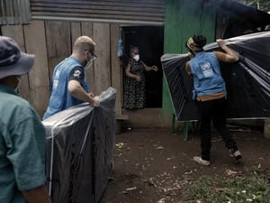 UNHCR staff deliver mattresses to a woman who stands in a doorway of a shelter.