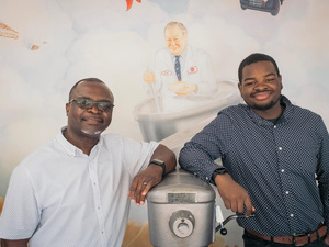 Two men stand either side of an industrial cake mixing machine in front of a mural-painted wall