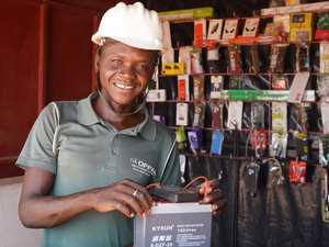 A man wearing a white hard hat tests a battery in a small electronics stall.
