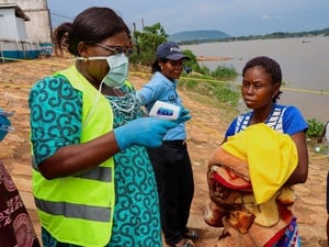 A woman wearing a mask prepares to take another woman's temperature next to a river, while a police officer stands nearby.