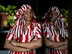 A woman in a red and white striped dress and headscarf leans against a window in which she is reflected